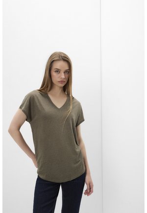 Jeune femme aux longs cheveux portant un t-shirt vert olive à col en V et manches courtes ainsi qu’un jean foncé, debout devant un fond blanc.