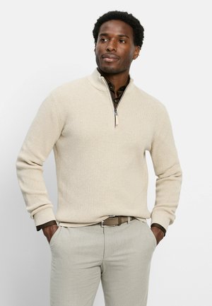Uomo con le mani nelle tasche che indossa un maglione beige con zip a quarto sopra una camicia scura e pantaloni chiari su uno sfondo semplice.