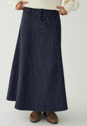 Longue jupe en jean foncé avec un détail à lacets sur le devant, portée avec un pull crème et des mocassins marron.