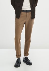 Mężczyzna w beżowych, podwijanych spodniach, białych skarpetkach, czarnych skórzanych butach, białej koszuli i ciemnobrązowej kurtce stoi na białej podłodze.