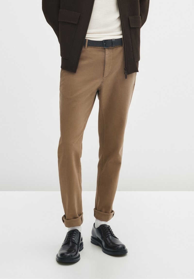 Mężczyzna w beżowych, podwijanych spodniach, białych skarpetkach, czarnych skórzanych butach, białej koszuli i ciemnobrązowej kurtce stoi na białej podłodze.