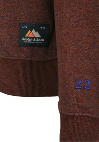 Suéter marrón rojizo con puños y dobladillo acanalados, que presenta una etiqueta negra con el logo "Scotch & Soda" y bordado azul "22" en la manga.