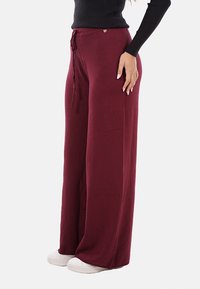 Pantaloni larghi color bordeaux realizzati in tessuto morbido, con vita regolabile e un piccolo dettaglio a forma di cuore argentato. Si abbinano a sneaker bianche.