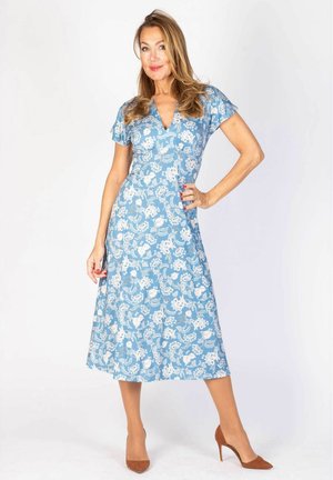 Nainen seisoo sinisessä kukallisessa midimekossa, jossa on lyhyet hihat ja V-aukko, ruskeat korkokengät jalassa, käsi lanteilla, valkoista taustaa vasten.