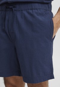 Marineblaue Shorts aus strukturiertem Stoff mit einem elastischen Bund und einer Kordel zum Verstellen. Sie verfügen über praktische seitliche Taschen.