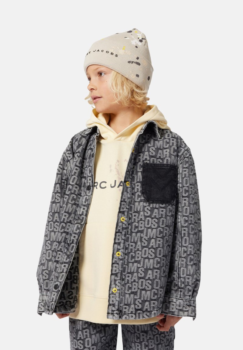 Grijze denimjack met herhaald logo patroon, zwarte pocketdetails, gele knopen, gedragen over een lichtgele hoodie, gecombineerd met bijpassende broek.