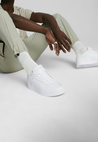 Baskets blanches avec une tige en cuir lisse, des lacets plats, une semelle légèrement surélevée et un logo sur le côté. Portées avec un pantalon et des chaussettes de couleur claire.