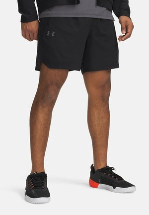 Mann trägt schwarze Sportshorts und schwarze Sneakers mit weißen Sohlen und roten Akzenten, steht auf weißem Hintergrund.