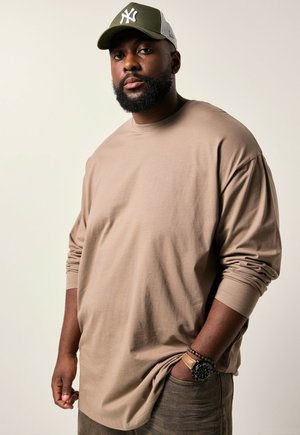 Beige langpoloshirt met een relaxte pasvorm, zachte textuur. Draag met een donkergrijze pet met een wit logo en aardetintenbroek.