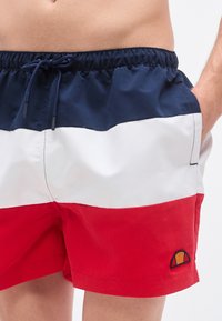 Μαγιό με μπλοκ χρωμάτων, με σκούρο μπλε ζώνη μέσης, λευκές και κόκκινες περιοχές, και κεντημένο λογότυπο στο αριστερό πόδι. Κατασκευασμένο από απαλό ύφασμα.