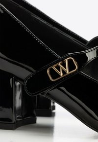 Zapatos Mary Jane de charol negro con tacones gruesos y un logo dorado "WD" en la correa.