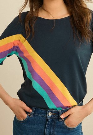 Femme portant une chemise bleu foncé avec des rayures diagonales arc-en-ciel en jaune, orange, violet et sarcelle, associée à un jean bleu, mains dans les poches.