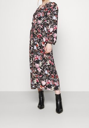 Kobieta w czarnej, kwiecistej sukience midi z długim rękawem i wiązaniem w talii, ubrana w czarne szpiczaste botki, stojąca na tle białego tła.