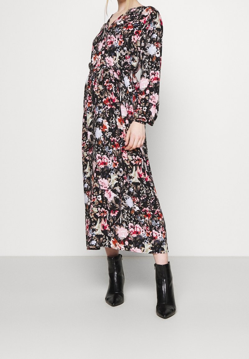 Femme portant une robe midi noire à manches longues avec des motifs floraux, une ceinture à la taille, et des bottines noires pointues, debout devant un fond blanc.