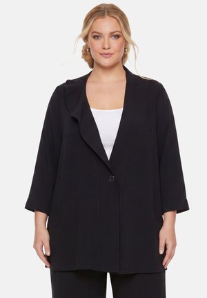 Frau mit blondem Haar trägt schwarzen offenen Blazer über weißem Oberteil, steht vor einem schlichten hellen Hintergrund und blickt nach vorne.