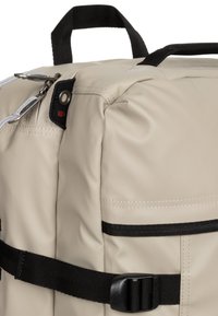 Beige ryggsäck med en strukturerad yta, svarta remmar, ett handtag på toppen och en kombination av dragkedjor och spännen för säker stängning.