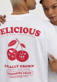 Deux personnes portent des t-shirts blancs ; le motif au dos montre des cerises rouges avec des visages et un texte faisant la promotion de fruits locaux et rafraîchissants.