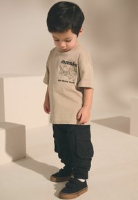 Beige bomulds t-shirt med sort grafisk tekst, parret med sorte cargo-bukser med flere lommer og sorte sneakers med velcro-stropper.