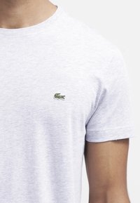 Camiseta gris jaspeada con cuello redondo, mangas cortas y un pequeño logotipo de cocodrilo bordado en verde en el lado izquierdo del pecho.
