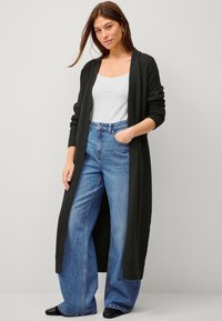 Schwarze Strickjacke mit langen Ärmeln und offenem Vorderteil, über einem weißen Tanktop und hoch taillierten blauen weit geschnittenen Jeans getragen.