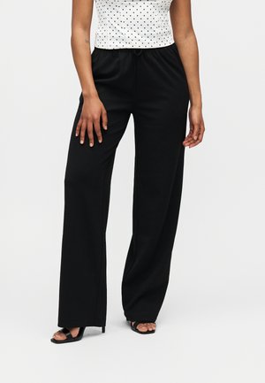 Kvinde iført sorte bukser med vide ben, sorte åbne hæle og en hvid ærmeløs top med sorte polkaprikker på hvid baggrund.