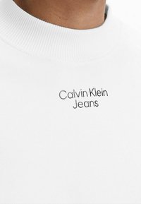 Sudadera blanca con cuello redondo acanalado y logotipo bordado "Calvin Klein Jeans" en negro. Tela suave con textura lisa.