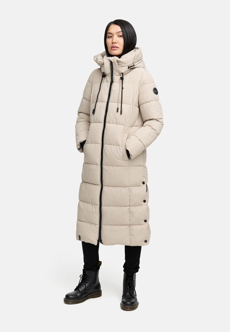 Cappotto lungo imbottito beige con cappuccio, chiusura frontale con zip, cordini e dettagli con bottoni. Indossato sopra pantaloni neri e stivali neri con lacci.