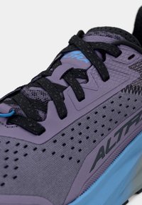 Zapatilla deportiva morada con cordones y detalles negros, presenta una parte superior texturizada, perforaciones para la transpirabilidad y una suela exterior azul.