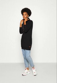 Vestido de moletom preto com capuz ajustável, punhos canelados, bolsos laterais e um patch de logotipo. Combinado com jeans azul claro e ténis brancos.