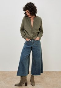 Olive green cropped bomber jacket with a zip front, paired with wide-leg blue denim jeans and gray suede ankle boots.