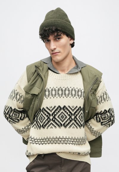 Μπεζ πλεκτό πουλόβερ με μαύρα γεωμετρικά σχέδια, φοριέται κάτω από ένα ελαιόχρωμο γιλέκο. Σκούρο πράσινο καπέλο από μάλλινη ύφανση από πάνω. Γκρίζο πουκάμισο με γιακά από κάτω.