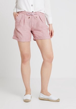 Vrouw met lichtroze shorts met trekkoord en opgerolde boorden, wit knopenoverhemd en witte instapschoenen, staand tegen een effen achtergrond.