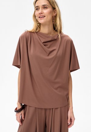 Kobieta w luźnej, brązowej bluzce z krótkim rękawem i dekoltem z marszczeniem oraz dopasowanych do niej szerokich spodniach, uzupełnionych kolczykami i bransoletkami.