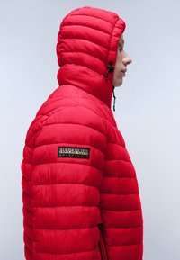 Rød pufferjakke med hætte, lodret quiltet mønster og lynlåslommer. Sidevisning viser et logo-patch på venstre ærme.