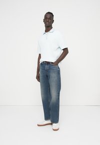 Φωτεινό γαλάζιο ριμπ πικέ πόλο μπλουζάκι, τζιν με ίσια γραμμή και ξεθωριασμένο χρώμα, λευκά αθλητικά παπούτσια με καφέ σόλες, σε όρθια στάση μπροστά σε λευκό φόντο.