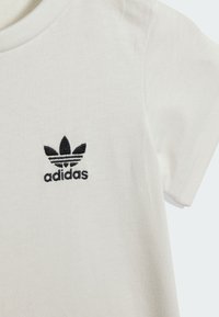 Biała bawełniana koszulka z okrągłym dekoltem. Na przodzie czarne haftowane logo Adidas z trzema liśćmi nad tekstem. Krótkie rękawy.
