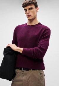 Gestrickter Rundhalspullover in tiefem Lila mit einem strukturierten Muster, kombiniert mit beigen Hosen und hält ein schwarzes Kleidungsstück.