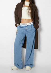 Donna con un lungo cappotto marrone, un maglione bianco corto, jeans larghi azzurri con una cintura bianca e sneaker bianche con piattaforma, con le mani nelle tasche.