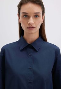 Camicia azzurra scuro con colletto a punta, texture liscia e chiusura frontale con bottoni. Nessun motivo o accento visibile.