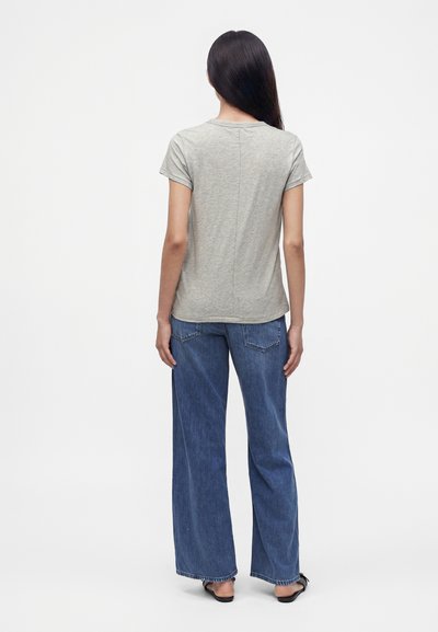 Femme debout de dos, portant un t-shirt gris clair à manches courtes, un jean bleu à jambes larges et des sandales noires ouvertes à l'avant sur fond blanc.