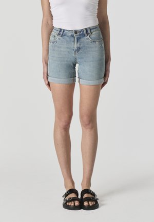 Mujer con pantalones cortos vaqueros de mezclilla azul claro con bordado frontal, combinados con sandalias negras con hebilla y una camiseta blanca sin mangas.