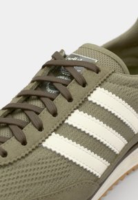 Olive green sneakers with three white stripes, brown laces, textured mesh upper, and rubber outsole. Brand logo on the tongue.