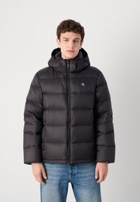 Czarna pikowana kurtka puchowa z zamkiem błyskawicznym i kapturem. Posiada pikowaną teksturę i małe logo na piersi. Noszona z niebieskimi dżinsami.