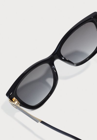 Schwarze, übergroße Sonnenbrille mit grauen Gläsern, glänzendem Kunststoffrahmen und goldenen Metallelementen an den Bügeln, mit einem eleganten Design.