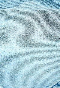 Tissu en denim bleu clair avec une texture subtile, présentant une surface lisse et un motif tissé, affichant de délicites coutures blanches le long des bords.