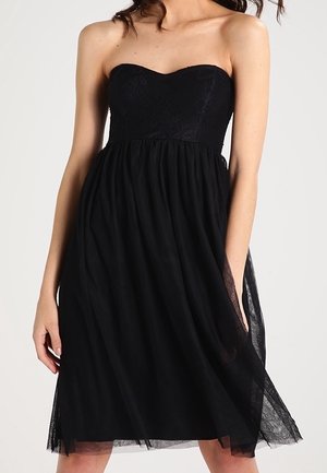 Vestido negro sin tirantes con corpiño de encaje y falda fluida y transparente, llevado por una mujer con el cabello suelto sobre un fondo liso.