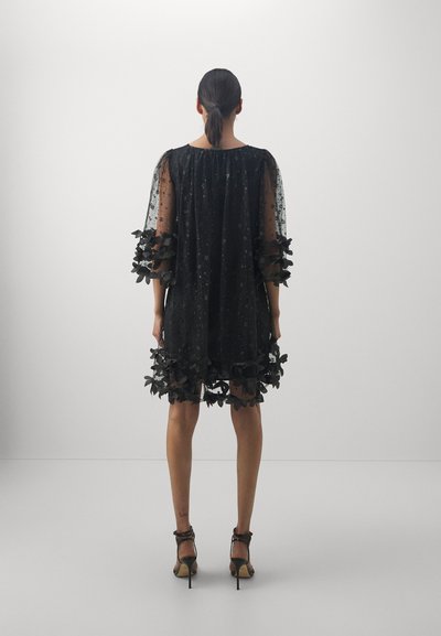 Rochie neagră din material transparent cu accente florale și mâneci bufante, prezentând un model cu stele și o margine ondulată. Pur tată cu pantofi cu toc.