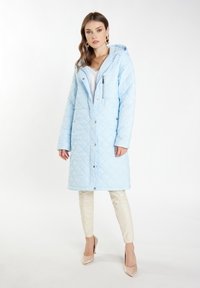Cappotto lungo trapuntato in blu chiaro, con cappuccio, tasca sul petto con zip e bottoni a pressione. Abbinato a pantaloni color crema e tacchi nudi.