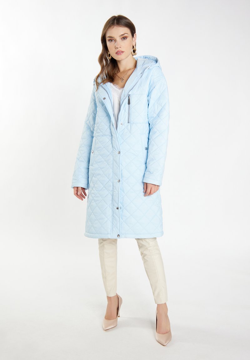 Cappotto lungo trapuntato in blu chiaro, con cappuccio, tasca sul petto con zip e bottoni a pressione. Abbinato a pantaloni color crema e tacchi nudi.