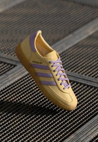 Geltonos sportinės bateliai su purpurinėmis akcentais, turintys tris juosteles, apvalų galiuką ir tekstūruotą pado dalį. Šoninėse dalyse matyti prekės ženklas "SPEZIAL."